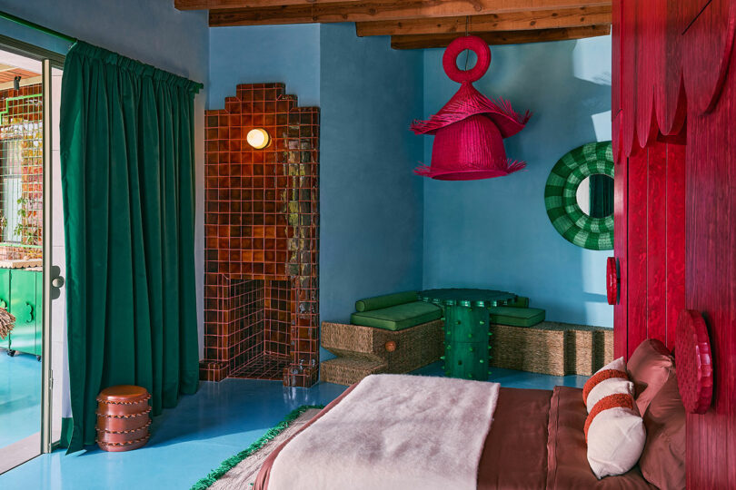 Colorful bedroom with blue walls, red accent decor, a bed with brown and white bedding, a green corner seating area, and a sliding glass door with green curtains.