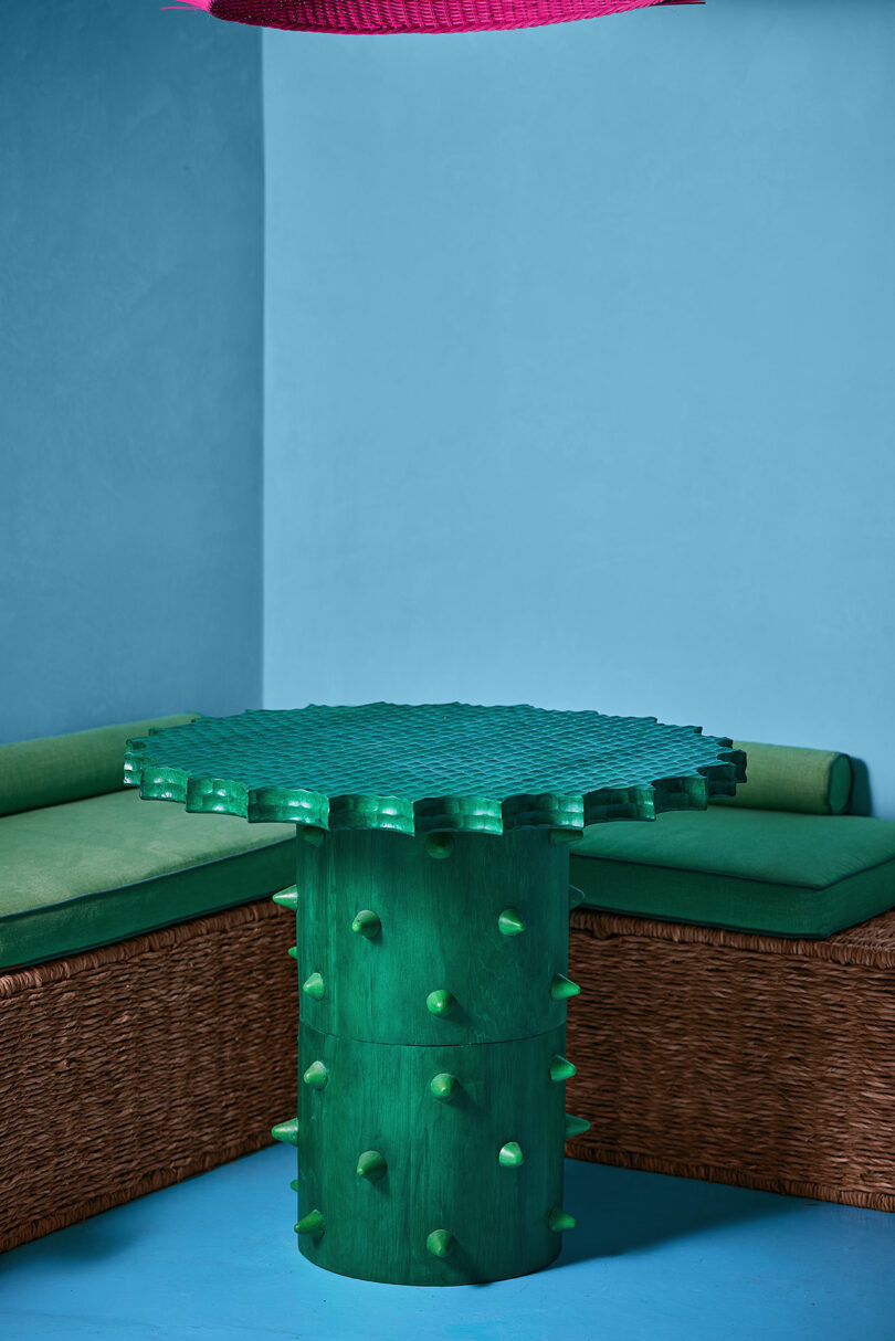 A green, textured table with knob-like protrusions stands in front of wicker seating with green cushions, against a blue wall.