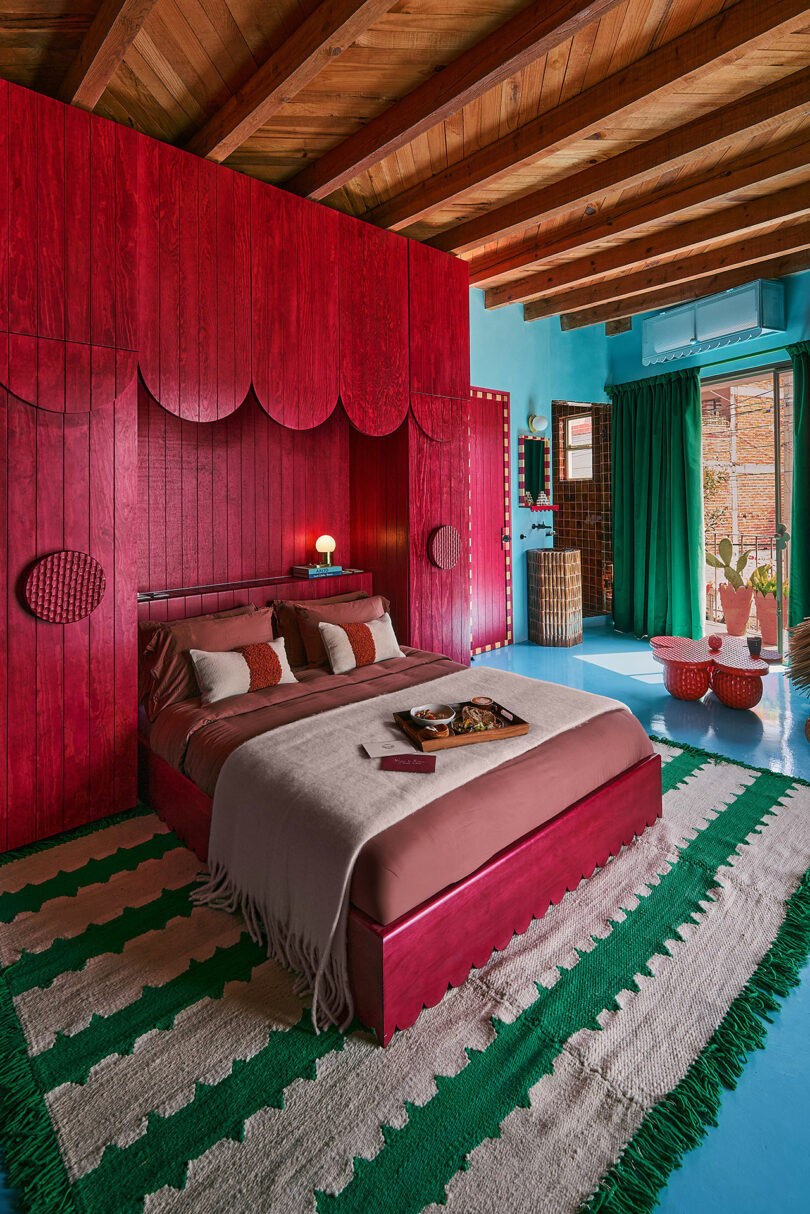 A bedroom with a bold red accent wall and bed, green curtains, a green and white striped rug, blue-painted floor, and wooden ceiling beams. Natural light enters through a window.
