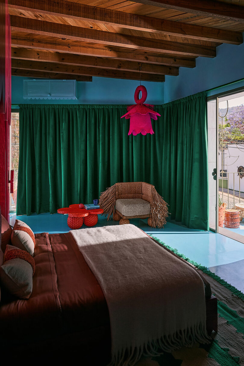 A bedroom with a wooden ceiling, green curtains, a brown bed, a unique pink ceiling lamp, a wicker chair, and red tables, with sunlight coming through a sliding glass door.