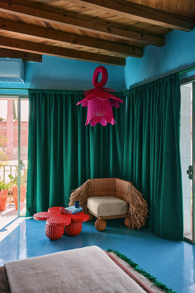 A room with blue walls and floor, green curtains, a woven chair with fringed arms, a red sculptural coffee table, and a large pink flower-shaped ceiling light.