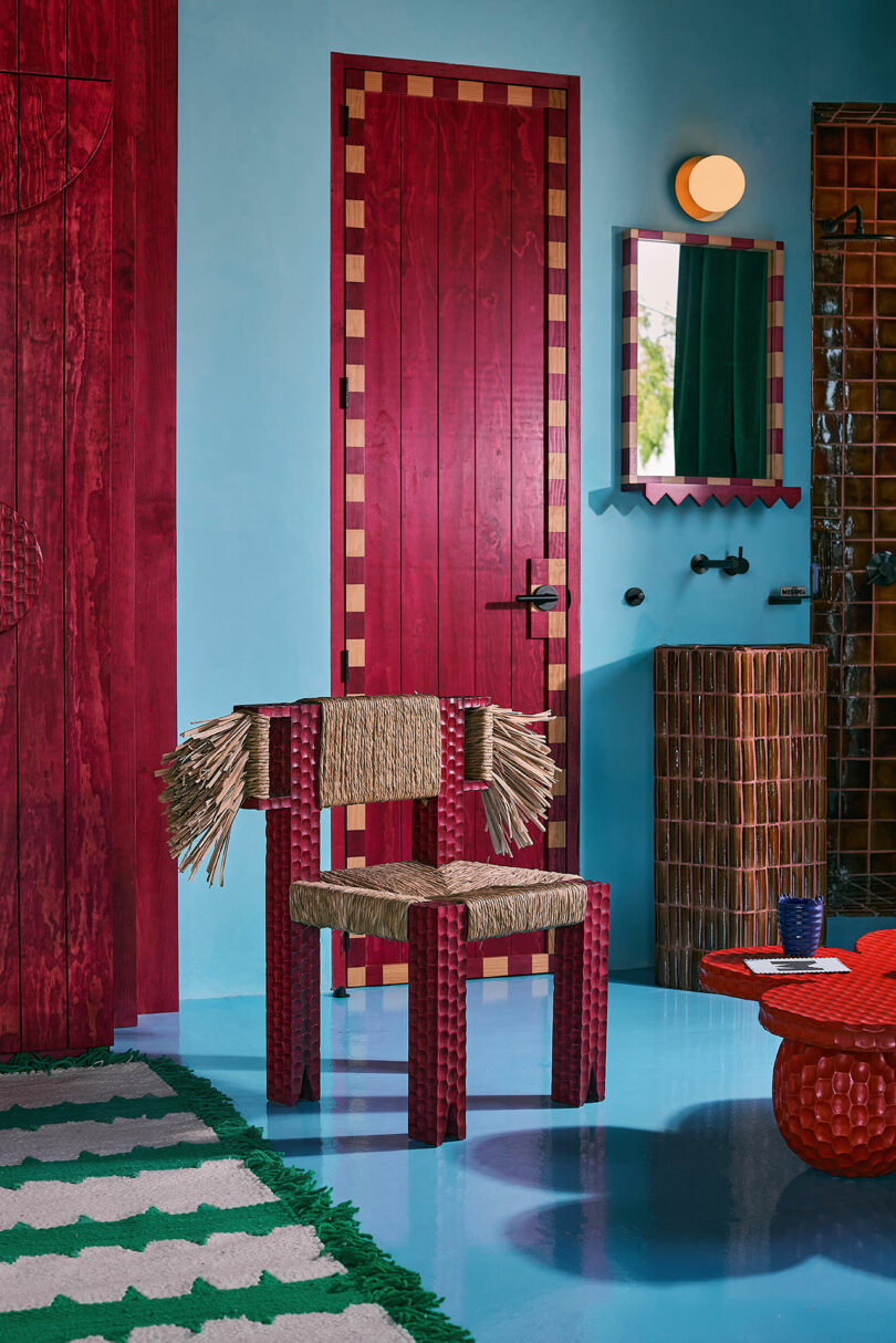 A room with blue walls, a red and tan checkered door, a woven chair with rope details, a round red table, a wall mirror, and a green-striped rug.