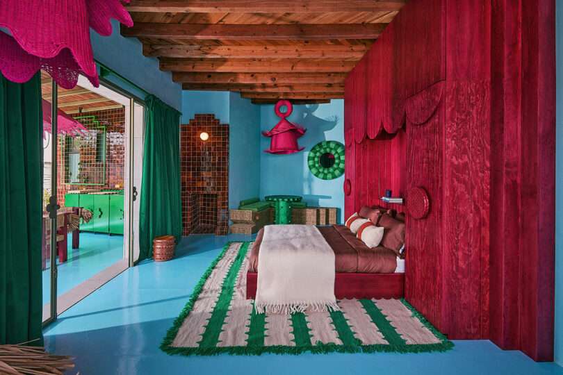 Bedroom with blue floor, red wood-paneled wall, green and white rug, brown bed, and open glass doors leading to an adjoining room with green decor and wood ceiling beams.