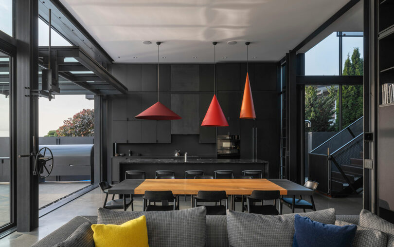 Modern kitchen and dining area with black cabinets, a wooden dining table, black chairs, and three geometric red pendant lights. Large windows provide natural light.