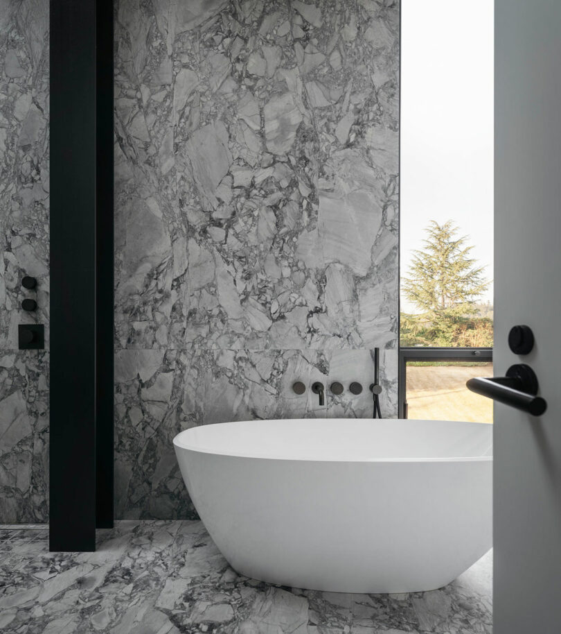 Modern bathroom with gray marble walls and floor, a freestanding white bathtub, black fixtures, and a window with a view of trees outside.