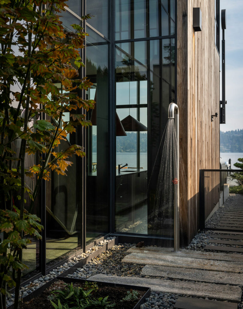 Outdoor shower with a curved metal fixture next to a modern glass and wood building, surrounded by gravel, stepping stones, and plants, with a view of a lake in the background.