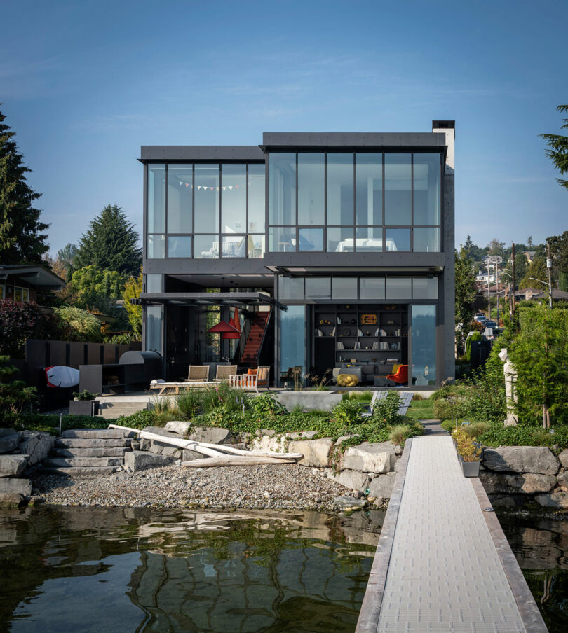 Modern two-story house with large glass windows, outdoor seating area, and a dock extending over the water, surrounded by greenery and stone landscaping.