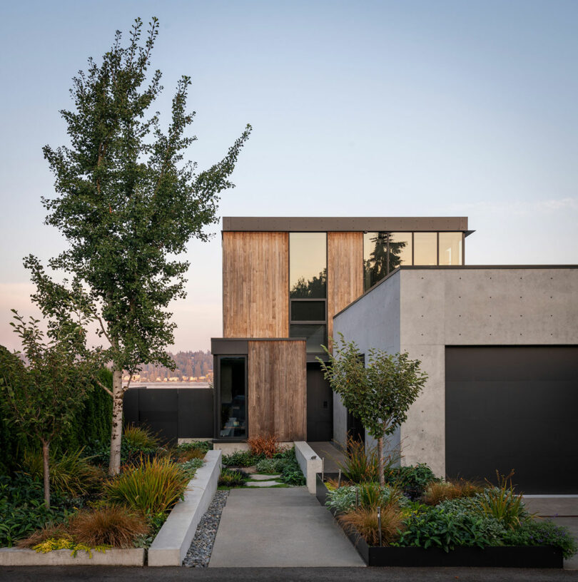A modern two-story house with wood and concrete exterior, large windows, and landscaped front yard with trees and plants.