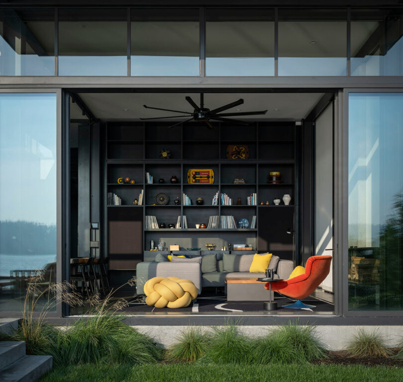 Modern living room with floor-to-ceiling windows, dark built-in shelves, colorful furniture, and a view of water and trees outside.