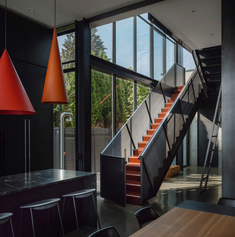 Modern interior with floor-to-ceiling windows, a black metal staircase with orange steps, red pendant lights, and minimalist black furniture.