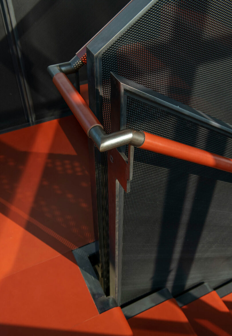 A close-up view of a modern metal staircase with red handrails and perforated metal panels, casting shadows on the red floor.