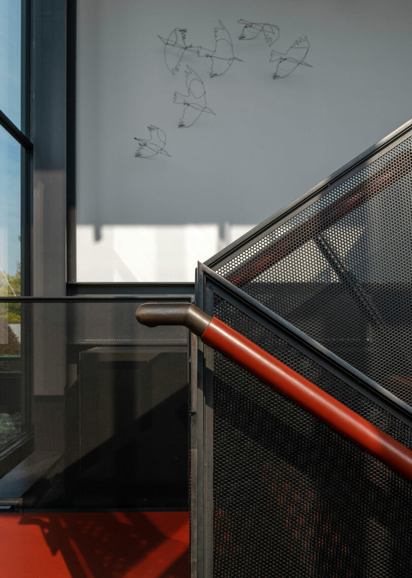 A staircase with a perforated metal railing and a red handrail, with a wall-mounted wire sculpture of birds above.