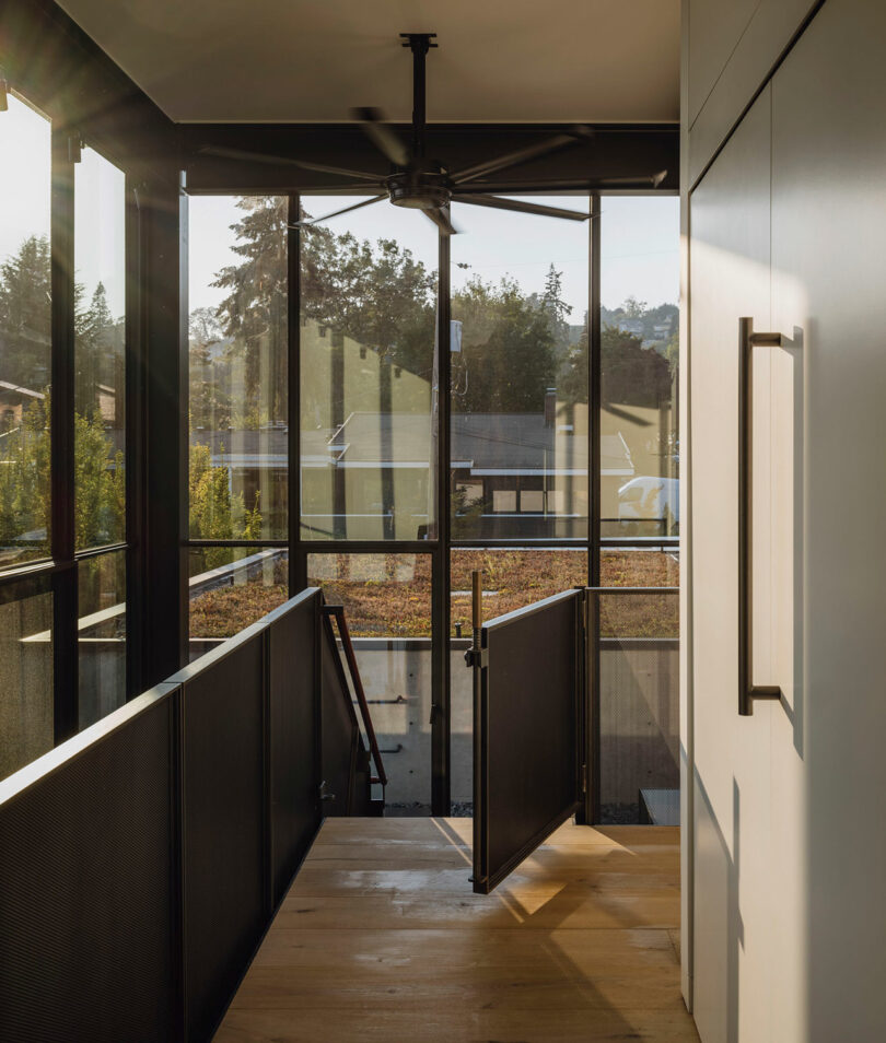 A modern stair landing with glass walls, black railings, a ceiling fan, and a view of trees and rooftops outside.