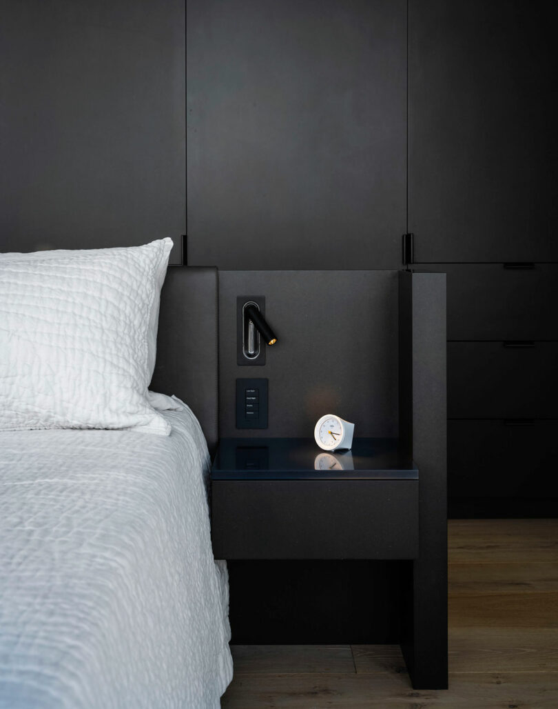 A modern black nightstand with a built-in reading light, electrical outlets, and a small white clock stands beside a bed with a white quilted pillow and blanket.