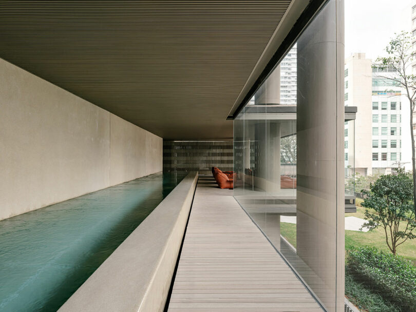 A modern covered walkway with a wooden floor runs beside a narrow indoor pool, separated from the outdoors by a large glass wall overlooking a landscaped area.