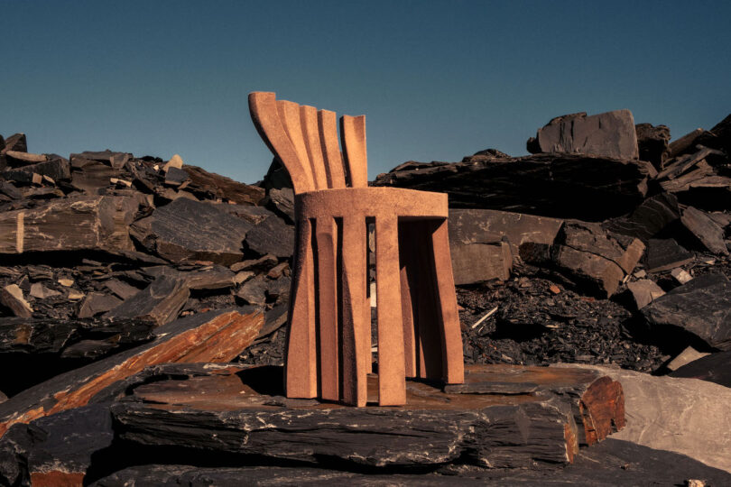 A sculptural wooden chair with curved slats stands on rocky terrain under a clear blue sky