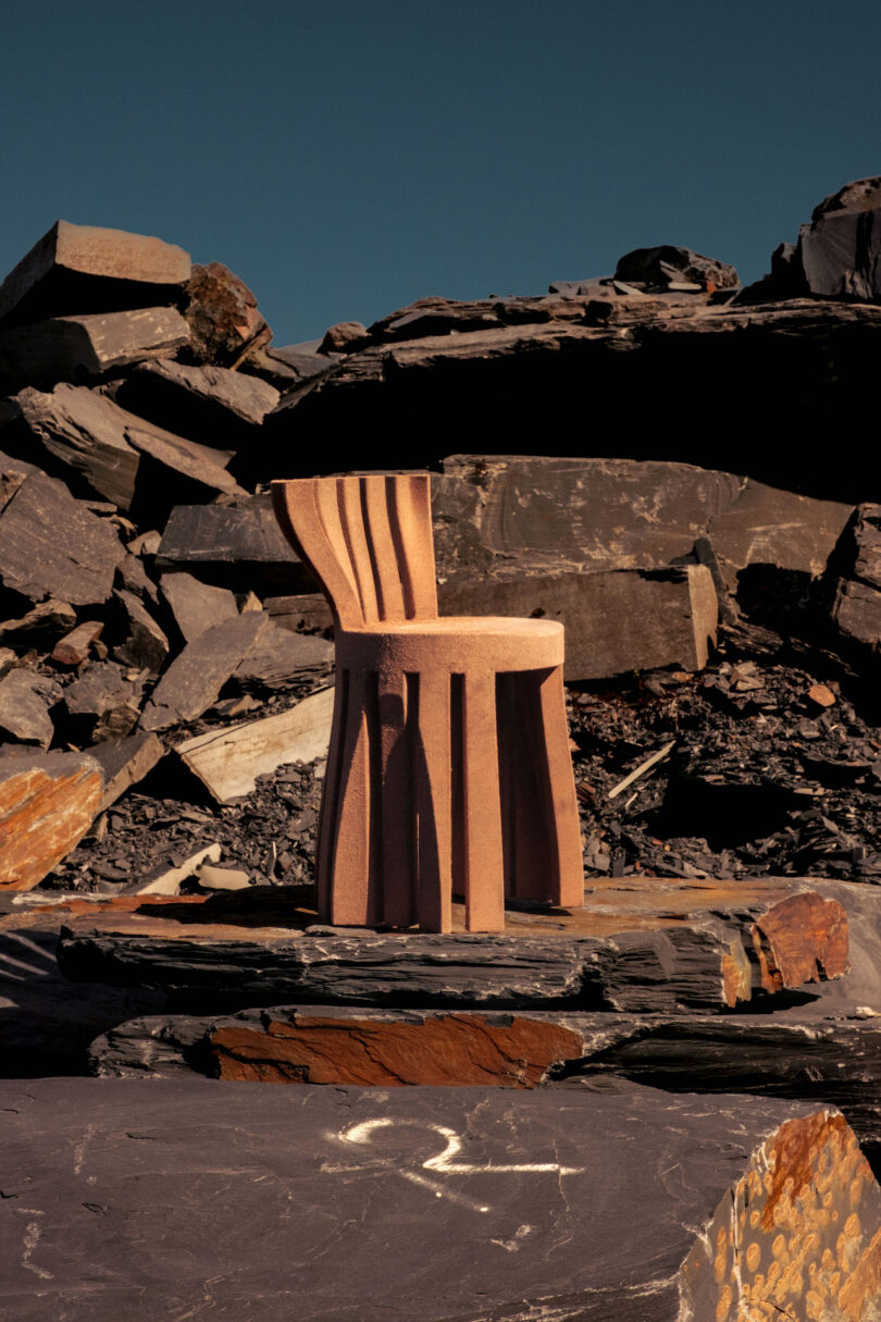 A sculptural wooden chair with curved slats stands on rocky terrain under a clear blue sky