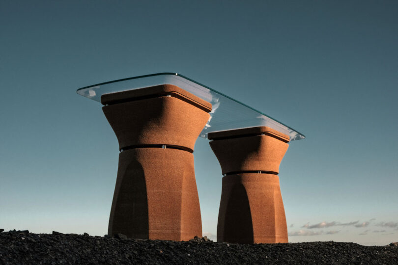 Two sculptural, hourglass-shaped brown bases support a rectangular glass tabletop, set outdoors on dark ground with a clear blue sky in the background