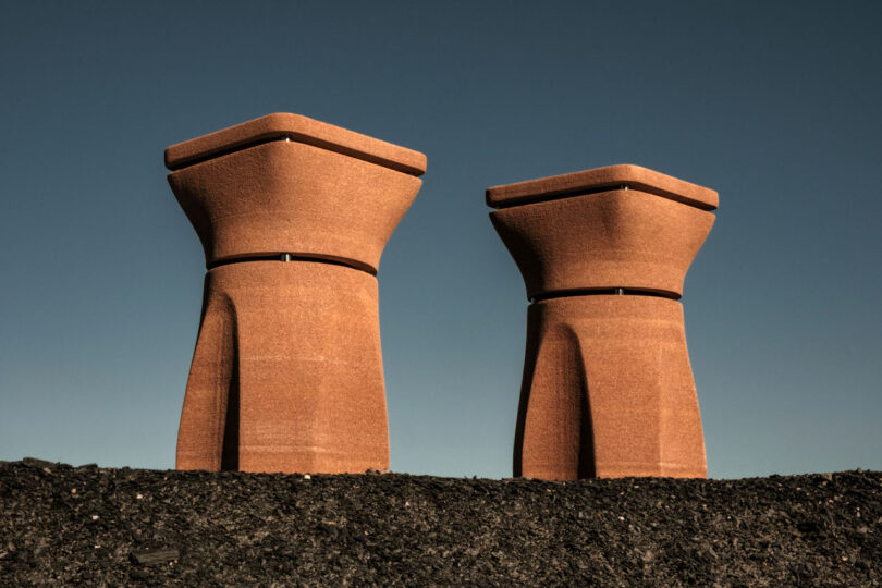 Two large, identical structures with a rounded, flared shape rise above a dark gravel surface against a clear blue sky