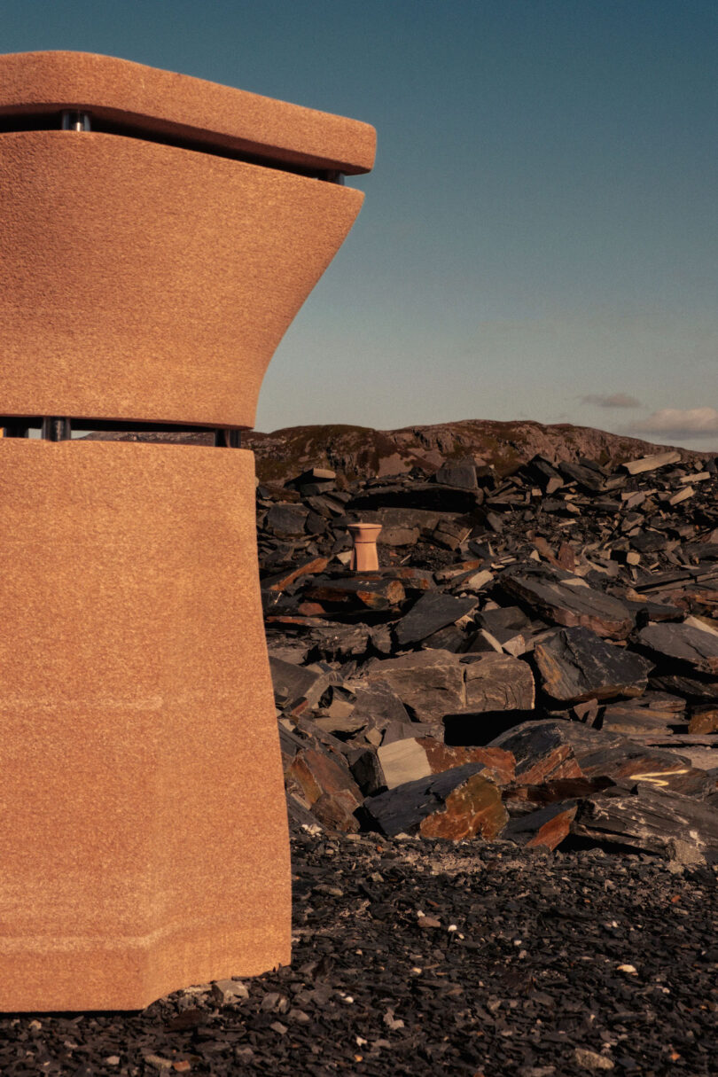 A large, rust-colored sculpture stands on rocky terrain, with a similar smaller sculpture visible in the background under a clear sky