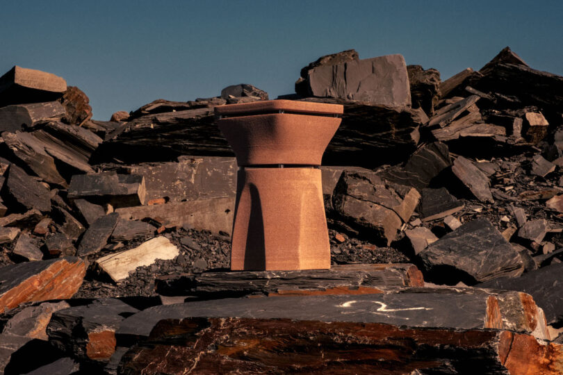A sculpted, geometric pedestal stands among jagged, dark slate rocks under a clear blue sky
