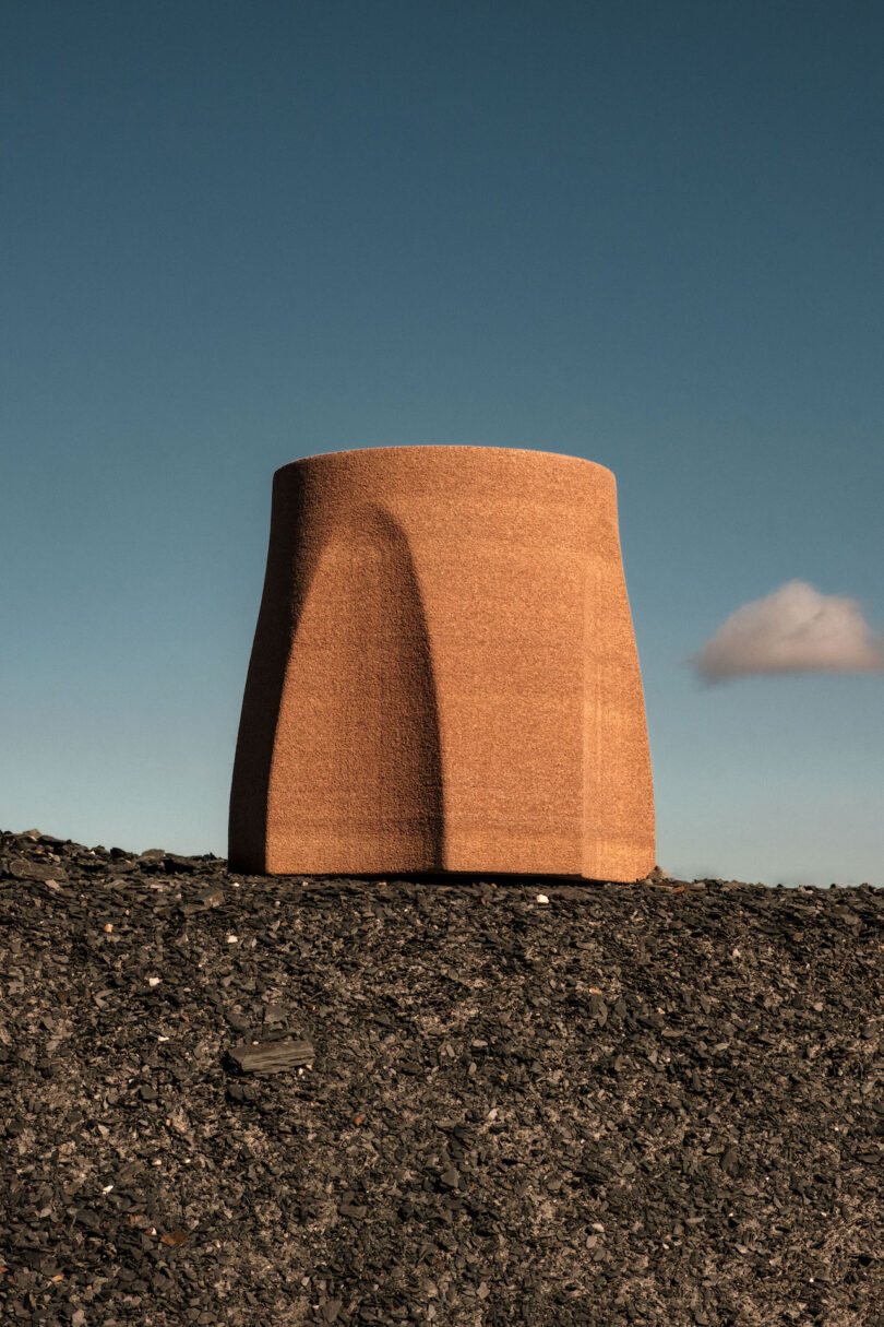 A large, cylindrical structure with smooth, curved surfaces stands on rocky ground under a clear blue sky with a single small cloud