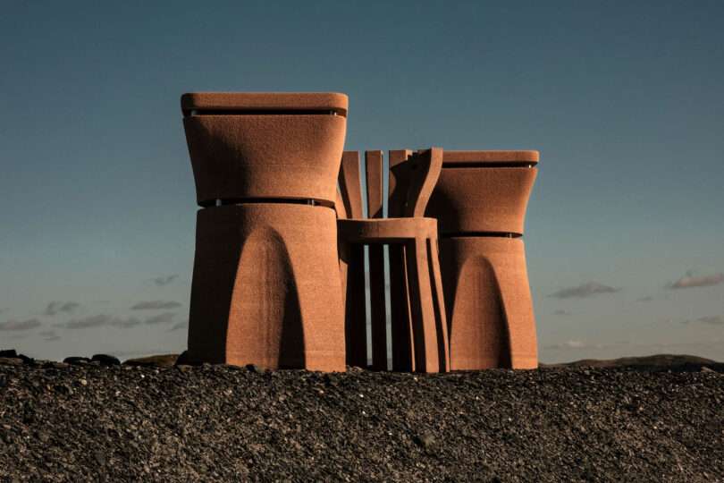 Three large, sculptural, rust-colored structures stand on a barren, rocky landscape under a clear sky