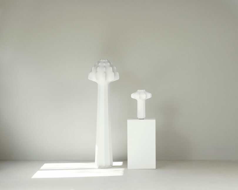 Two white sculptural lamps, one tall and one short, stand against a plain light gray wall; the shorter object is placed on a white pedestal.
