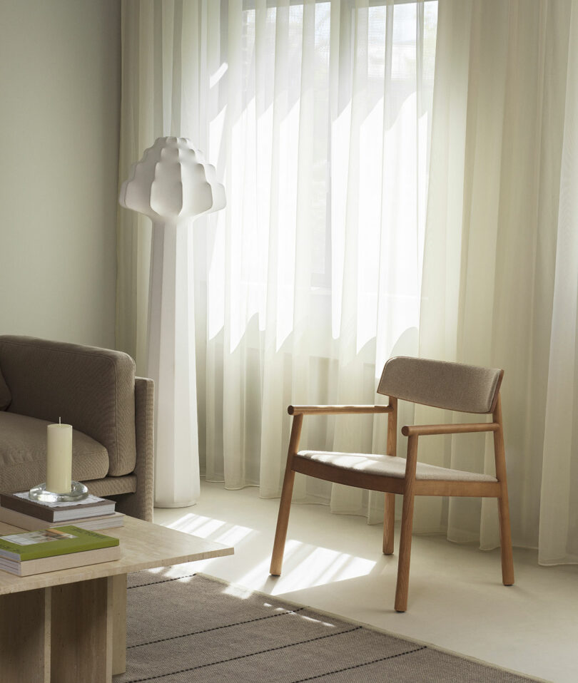 A minimalist living room with a beige sofa, wooden chair, tall white floor lamp, light curtains, and a small coffee table with a candle and books.