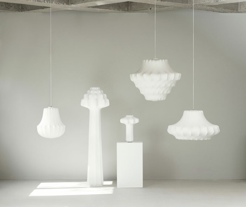 A minimalist room with three white pendant lamps and two sculptural white floor lamps against a plain gray wall.