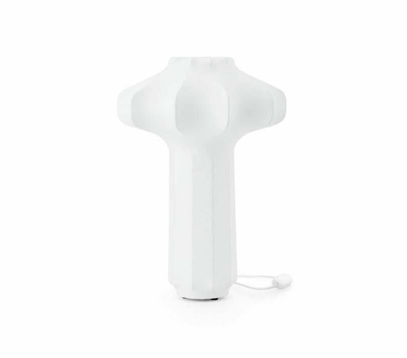 A white, modern, sculptural lamp with a rounded top and vertical grooves, featuring a pull cord at the base, set against a plain white background.