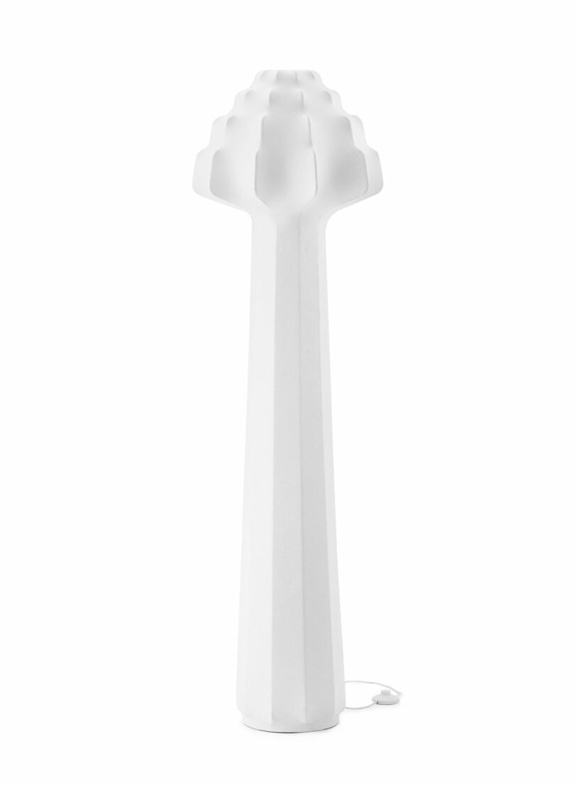 Tall, white, sculptural floor lamp with a faceted base and a stepped, rounded top; electrical cord visible at the base.