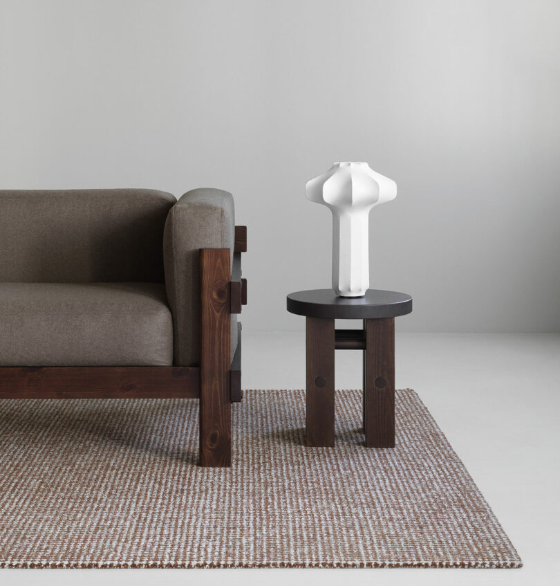 A brown wood-framed sofa sits on a woven rug next to a small round side table with a modern white sculptural vase on top, against a plain light-colored wall.