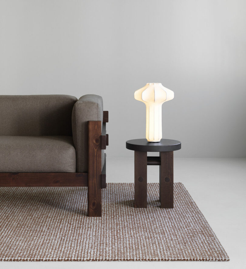 A brown wooden sofa with grey cushions is next to a round black side table holding a white sculptural lamp, all on a textured area rug in a minimal setting.