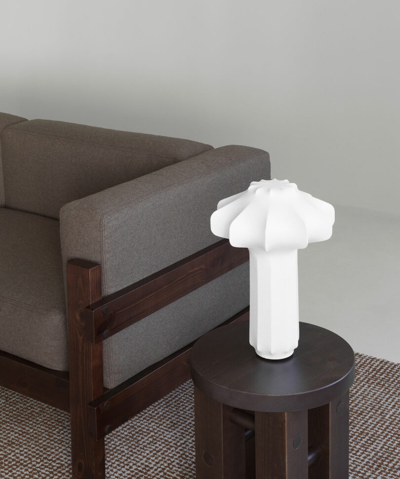 A modern white sculptural lamp sits on a round dark wood side table next to a brown upholstered sofa on a woven rug in a minimalist room.