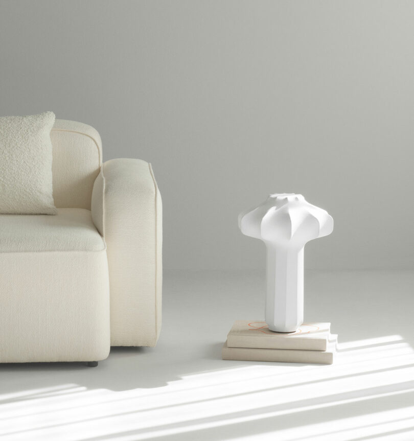 A modern white sculptural vase sits on stacked books next to an off-white sofa in a minimal, sunlit room.