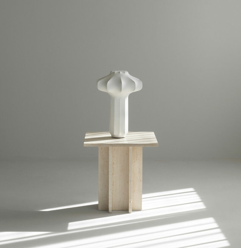 A sculptural white vase with geometric curves sits on a minimalist square stone pedestal in a softly lit, neutral-toned room.