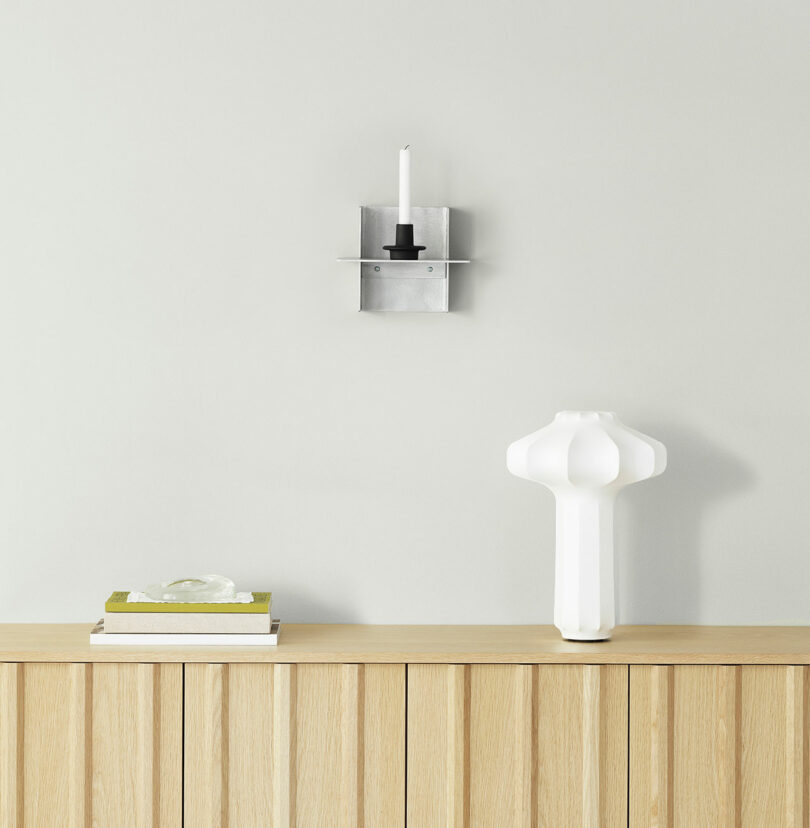 A wooden cabinet with a white sculptural lamp, a closed book, and a small decorative object on top; above is a wall-mounted metal candle holder with a white candle.