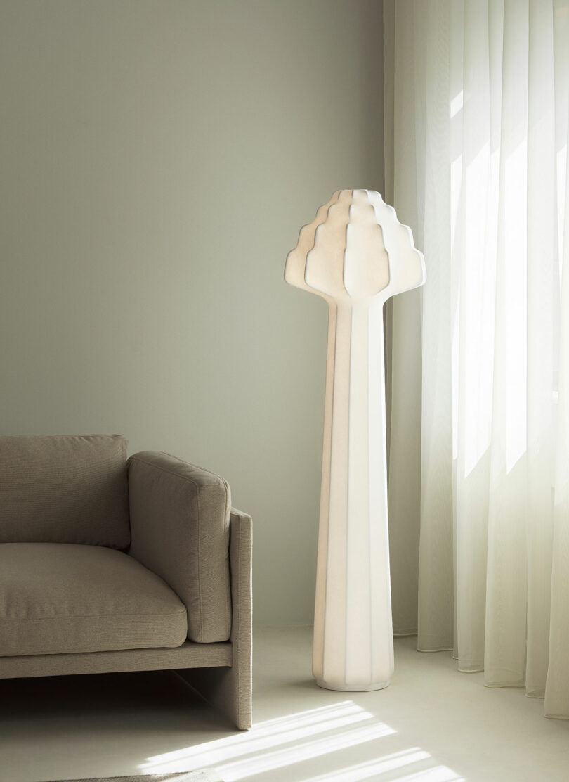 A tall, sculptural white floor lamp stands beside a light beige sofa in a minimal, sunlit room with sheer curtains.