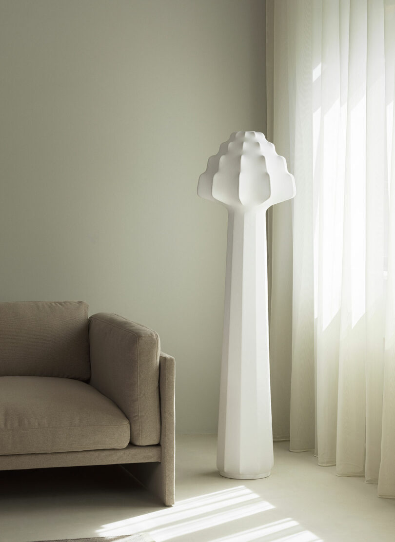 A minimalist room with a beige sofa, sheer white curtains, and a tall, sculptural white floor lamp against a light-colored wall.