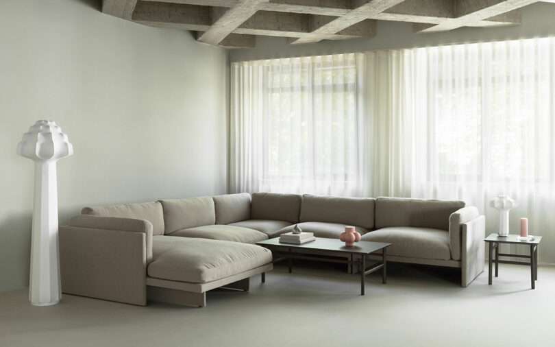 A modern living room with a beige sectional sofa, two coffee tables, a white sculptural floor lamp, a small table lamp, and sheer curtains over large windows.