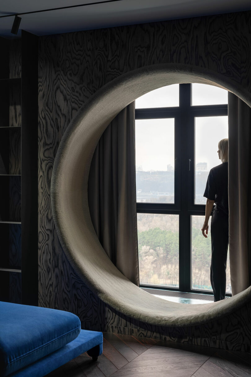 Person standing by a large window with an oval-shaped frame, looking outside at a landscape of trees and distant buildings, in a modern room with dark walls and blue furniture.