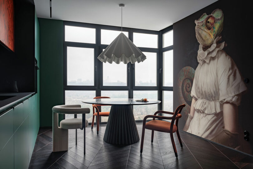 Modern dining area with large windows, a round table, mixed chairs, and a wall mural of a chameleon wearing a ruffled collar and dress.