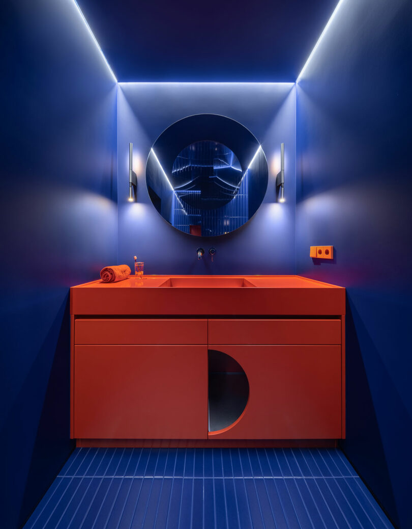 A modern bathroom with blue walls and floor, an orange vanity with a circular cutout, a round mirror, LED strip lighting, and two wall sconces.