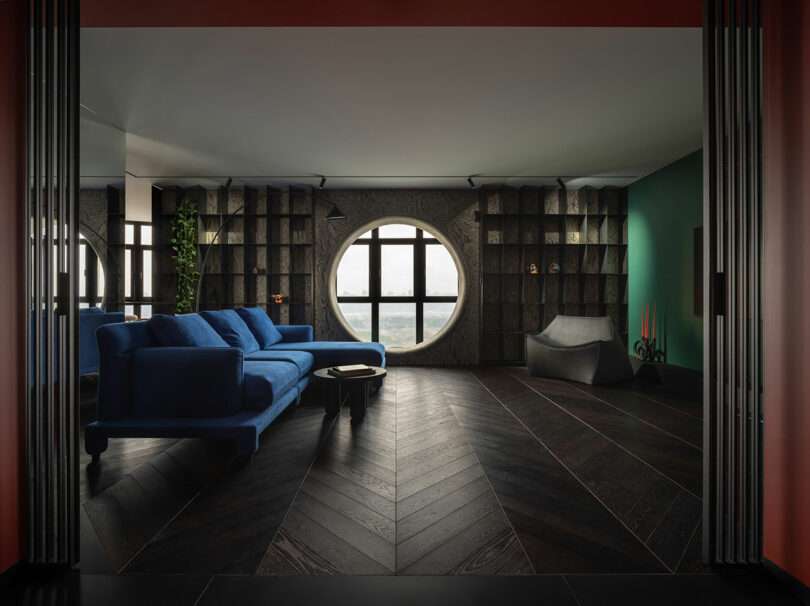 Modern living room with a blue sofa, circular window, dark wood floors, built-in shelving, and green accent wall. Sculptural gray chair and candles add decorative touches.