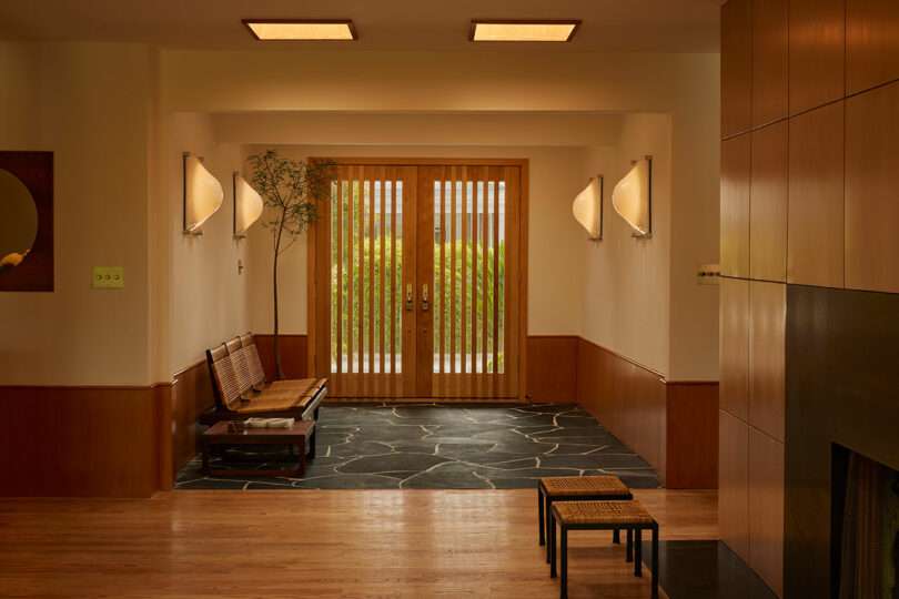 Entryway with wooden double doors, a bench with cushions, stone tile flooring, wall sconces, and a small plant in the corner. Light wood paneling and flooring throughout.