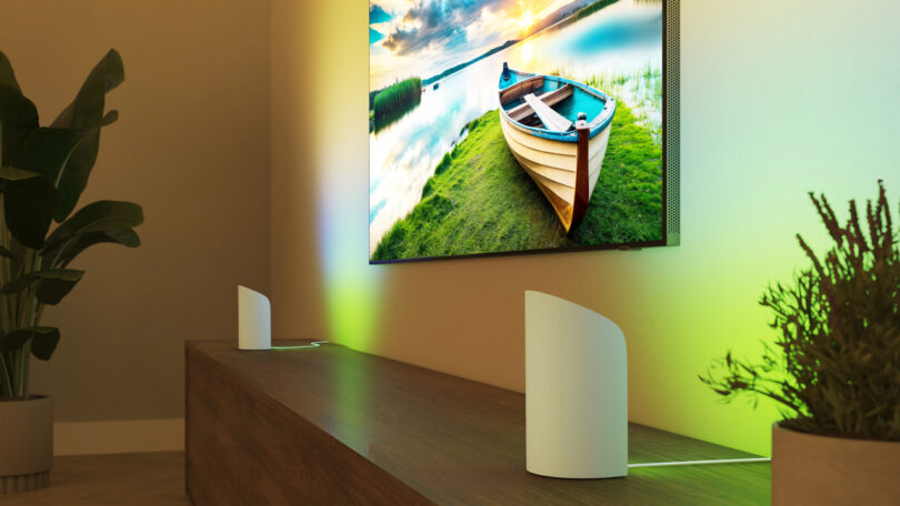 A modern living room with a large wall-mounted TV displaying a boat on water, surrounded by ambient colored lighting, plants, and two white electronic devices on a wooden cabinet.
