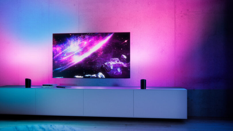 A flat-screen TV displaying a space scene is mounted on a pink-lit wall above a white media console with two speakers and some items.