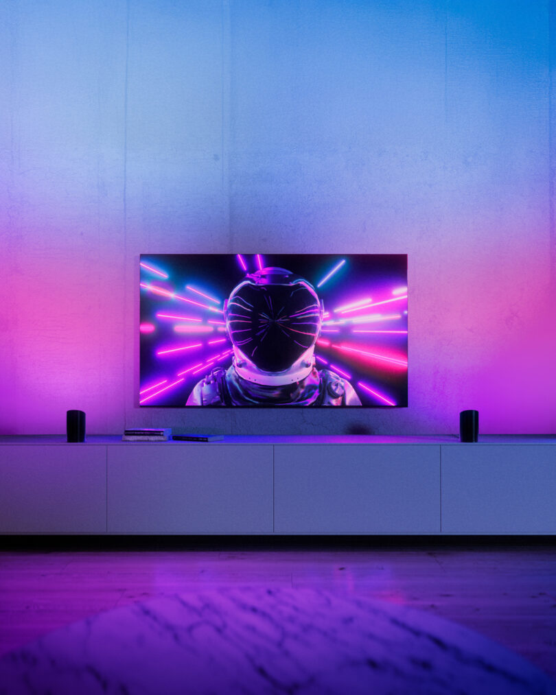 A modern living room with a TV on a white cabinet displaying a person in a dark helmet and suit, surrounded by colorful neon lights. The room is lit with purple and blue ambient lighting.