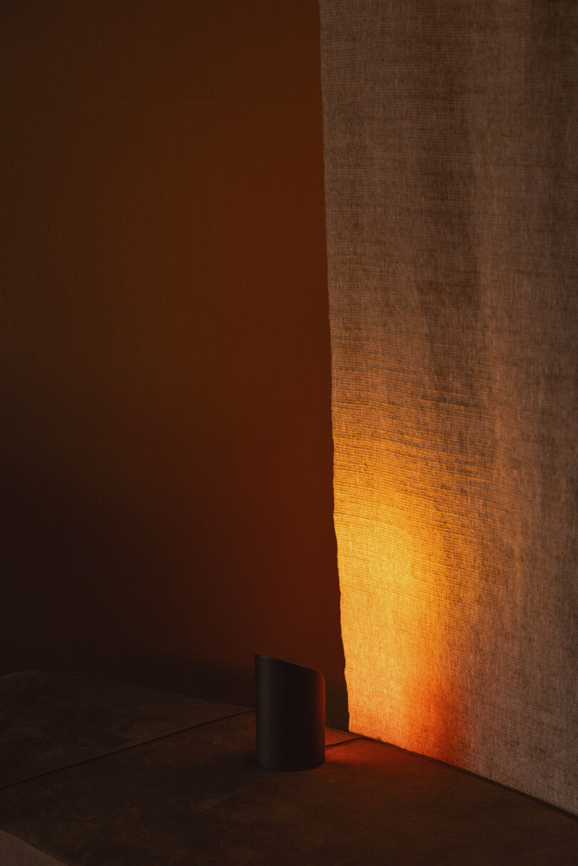 A warm light illuminates a beige fabric curtain, casting an orange glow on the wall and surface with a black cylindrical object in the foreground.